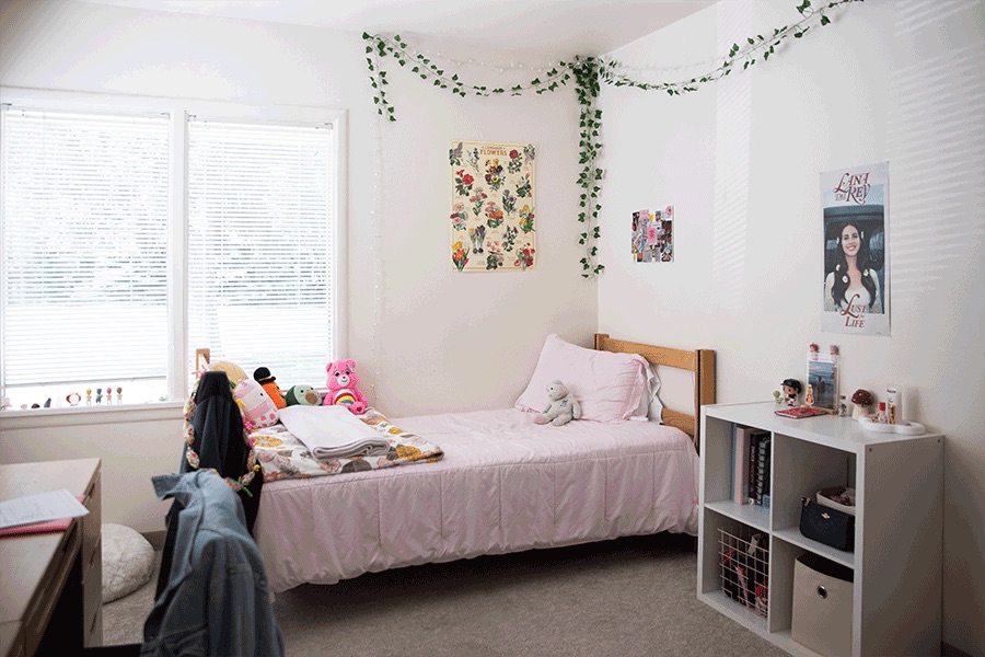 A cozy bedroom with a single bed, bookshelf, desk, large window with blinds and various decor.
